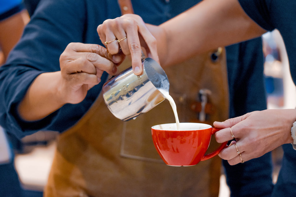 Person pouring milk from a pitcher into a red cup held by another person.